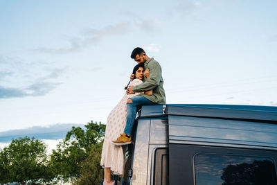 Side view of young man sitting on camper van roof and embracing girlfriend while chilling together in summer evening in countryside