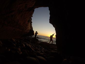 Silhouette couple standing by cave in sea against sky during sunset