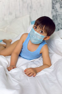 Boy in a blue t-shirt and a medical mask sits on a bed in quarantine during the covid-19 epidemic