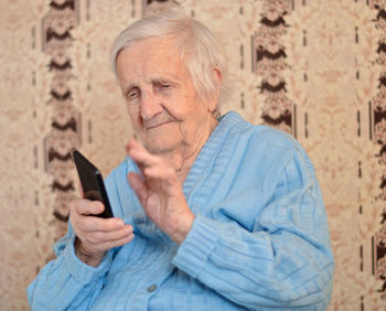 Happy elderly 90-year-old woman with glasses wearing a blue jacket smiles using a smartphone.