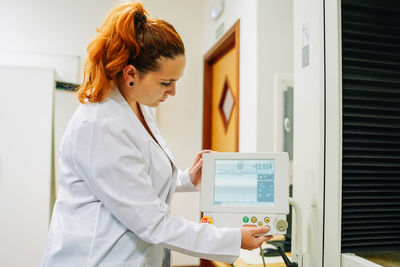 Serious female scientist pressing button of modern electronic machine with display while setting up equipment during work in light lab