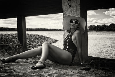 A woman in a swimsuit, hat and sunglasses sits on the sand on the riverbank at a concrete pier