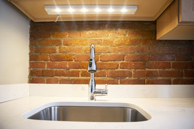 Closeup of elegant, silver kitchen sink with faucet, white counter top, led light and red brick wall