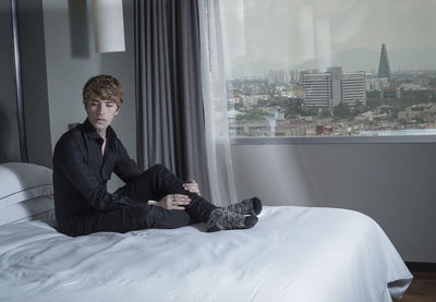 Young man sitting on bed against window