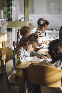 Girl writing on paper while sitting with students at bench in classroom