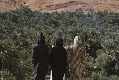 Rear view of friends wearing djellabas while standing against palm trees at desert
