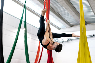 Bearded adult dancer in black leotard hanging upside down on aerial silk during rehearsal in studio