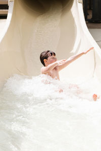 Full body of excited shirtless little boy with wet dark hair riding on water slide while spending sunny summer day in aqua park