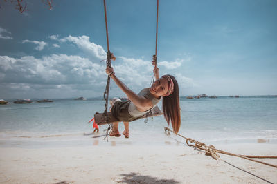 Portrait of smiling young woman sitting on swing at beach against sky