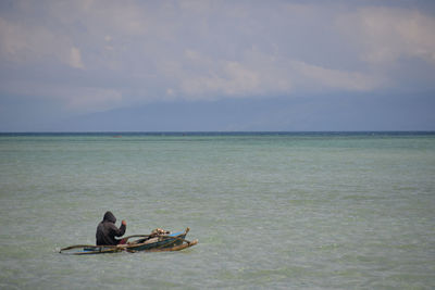 Rear view of man sitting in boat on sea against cloudy sky
