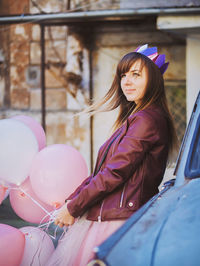 Young woman holding pink balloons while leaning on car