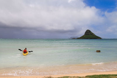 Rear view of people canoeing in sea against cloudy sky