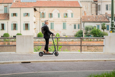 Side view full length young businessman in a suit riding an electric scooter while commuting to work 