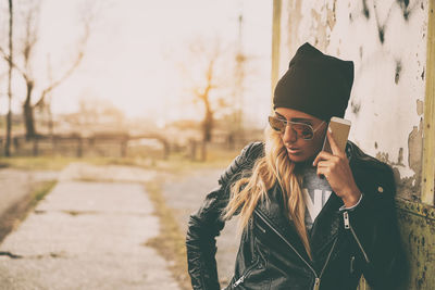 Fashionable young woman wearing leather jacket and sunglasses using smart phone against wall