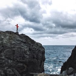 Man with arms outstretched on rock formation by sea against cloudy sky