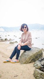 Portrait of woman wearing sunglasses sitting on rock at beach against sky