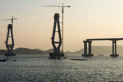 Incomplete bridge columns by cranes amidst sea against sky during sunset