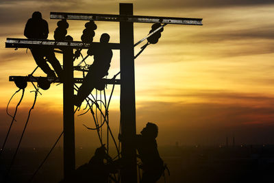 Silhouette people working on telephone pole against sky during sunset