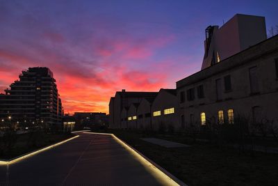 Road by illuminated buildings against sky at sunset