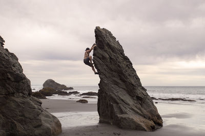 Side view of man climbing rock formation against cloudy sky