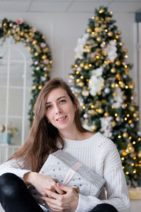 Girl in a white sweater sits with a christmas present on the background of a christmas tree. 