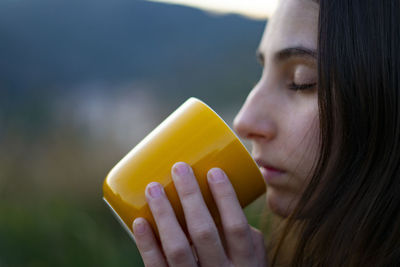 Young woman drinking and smelling coffee in a yellow cup in the woods.