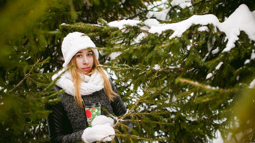 Young woman wearing warm clothing while holding disposable cup amidst trees during winter
