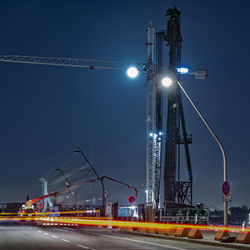 Light trail on street by crane against sky at night