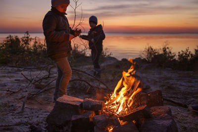 People standing by bonfire against sky during sunset