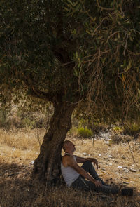 Adult man in white tank top and jeans taking rest under olive tree in summer