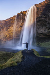 Full length of hiker with arms outstretched standing against waterfall
