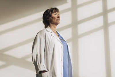Low angle view of female doctor with hand in pocket standing against wall at hospital