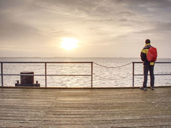 Rear view of man standing on railing against sea during sunset