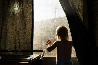 Rear view of boy looking through window while standing at home