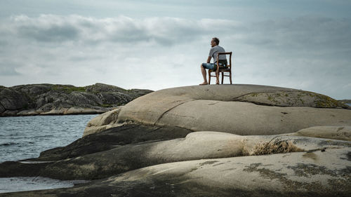 Rear view of man sitting on rock by sea against sky