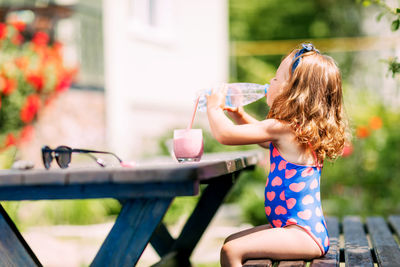 A little girl drinks water from a plastic bottle, sits on a bench in the garden on a hot summer day