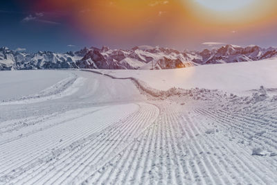 Tire tracks on snow landscape against sky during sunset