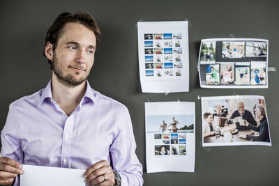 Mid adult businessman holding paper while standing against wall in office