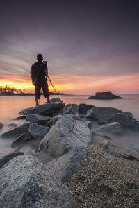 Man with tripod standing on rock at beach against sky during sunset