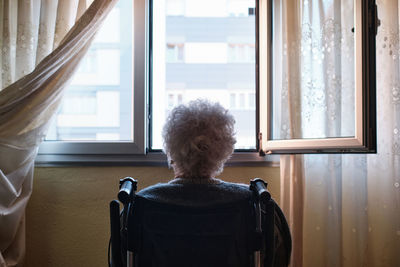 Back view of anonymous thoughtful senior disabled female in wheelchair sitting near opened window with curtains in light room at home