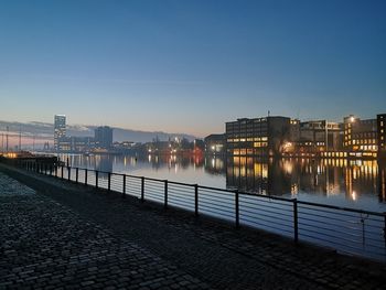 Illuminated city by river against sky at dusk