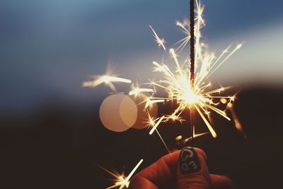 Cropped hand of woman holding sparkler against sky at dusk