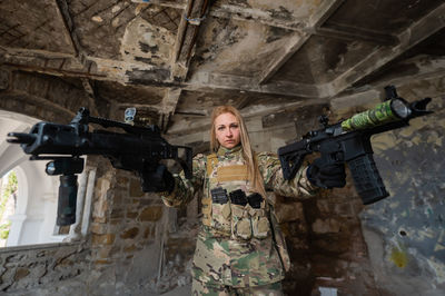 A woman in an army uniform aims to shoot a firearm in an abandoned building. 