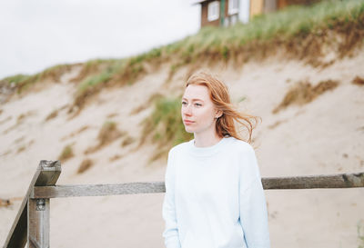 Portrait of young red haired woman in light blue sweater on sand beach by sea in storm