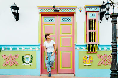 Full body of female traveler standing near residential building with multicolored door and creative decorated plinths during trip in guatape city