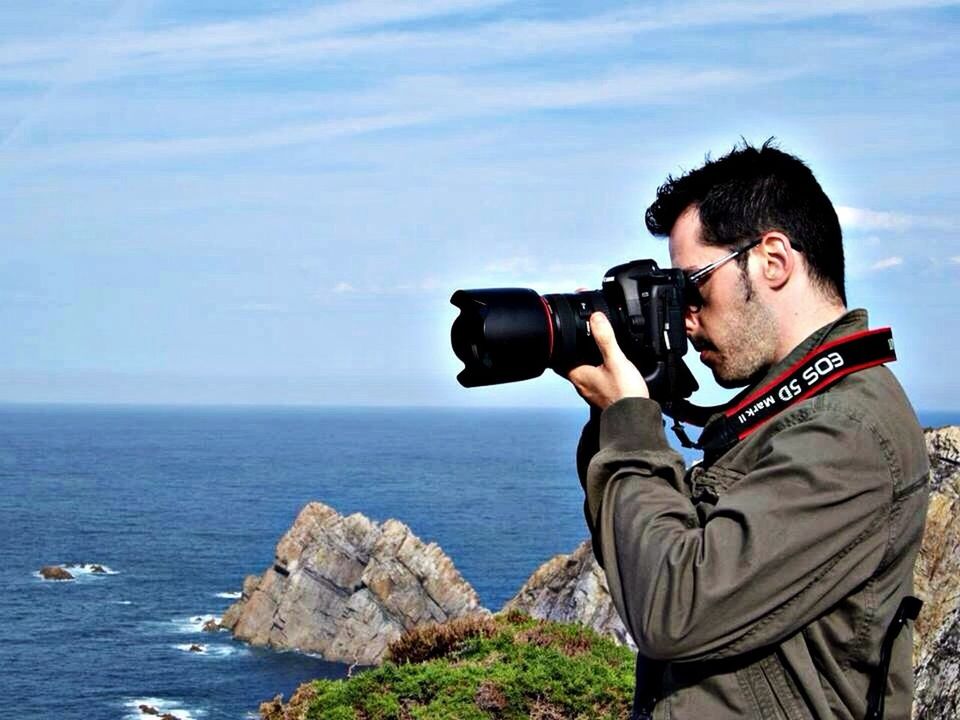 lifestyles, sea, leisure activity, young adult, water, sky, young men, photography themes, photographing, casual clothing, sitting, technology, person, waist up, horizon over water, rock - object, standing, three quarter length