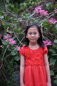 Portrait of cute girl wearing red dress while standing against flowering plants