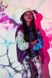 Young dominican female teenager in trendy outfit and hat standing near white wall with creative abstract projections and looking at camera while with arms crossed