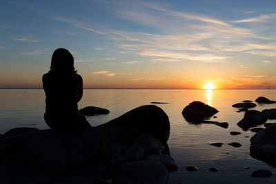 Rear view of silhouette woman sitting on rock at beach during sunset