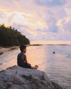 Man sitting on rock looking at sea shore against sky